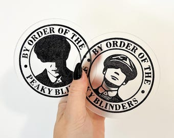 Könnte beinhalten: Zwei runde, transparente Acryl-Untersetzer mit schwarzen Druckdesigns. Die Untersetzer zeigen den Text "By Order of the Peaky Blinders" und eine Silhouette eines Mannes mit einer Mütze.