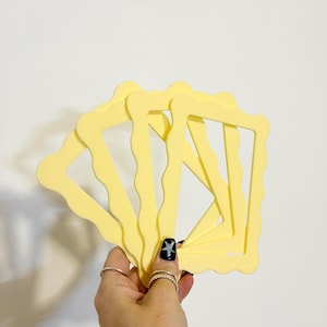 May include: A set of four pale yellow rectangular frames with wavy, scalloped edges. The frames are arranged in a fanned-out, overlapping position, showcasing their uniform light colour and consistent, decorative, undulating border design.