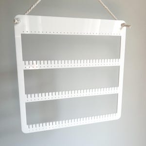 May include: White acrylic earring holder with three rows of holes for storing earrings. The holder is hanging from a white rope.