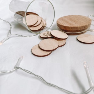 K&ouml;nnte beinhalten: Ein Glasgef&auml;&szlig; mit einem Holzdeckel, gef&uuml;llt mit kleinen Holzscheiben. Die Scheiben tragen die Worte "You are loved" aufgedruckt. Das Gef&auml;&szlig; steht auf einer wei&szlig;en Oberfl&auml;che mit einer Lichterkette.