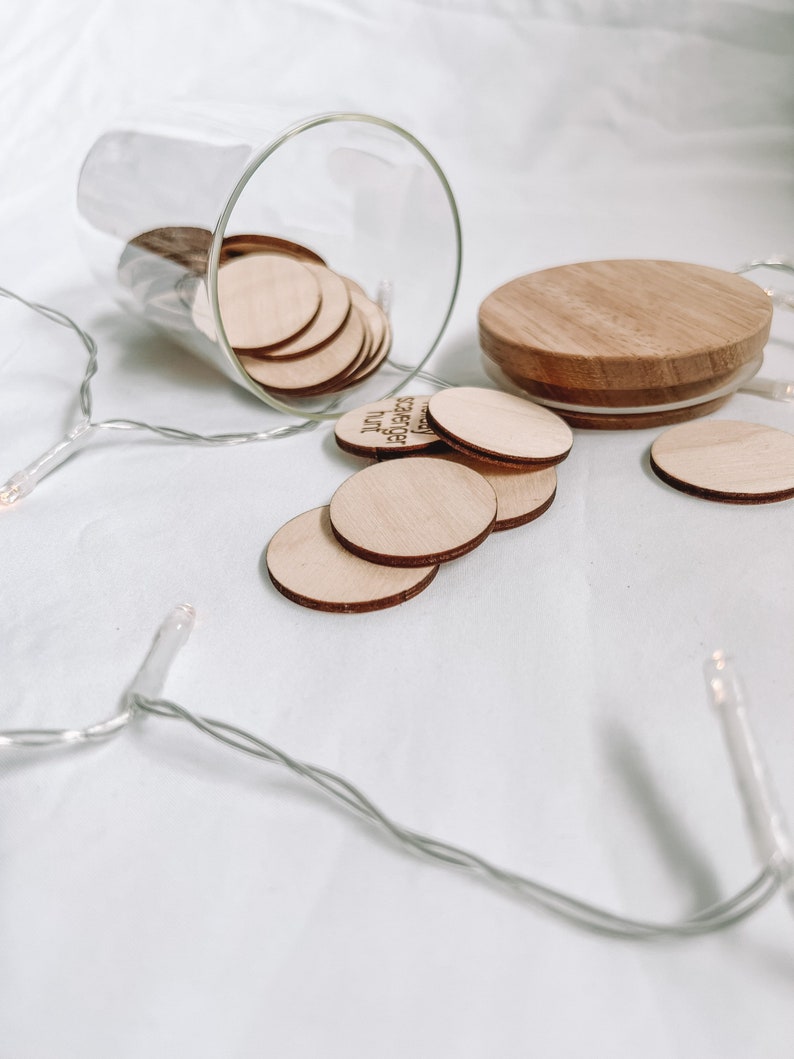 K&ouml;nnte beinhalten: Ein Glasgef&auml;&szlig; mit einem Holzdeckel, gef&uuml;llt mit kleinen Holzscheiben. Die Scheiben tragen die Worte "You are loved" aufgedruckt. Das Gef&auml;&szlig; steht auf einer wei&szlig;en Oberfl&auml;che mit einer Lichterkette.