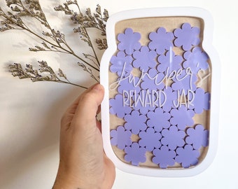 Könnte beinhalten: Ein weißer Holztopf mit einer durchsichtigen Vorderseite, gefüllt mit lila Blüten-förmigen Spielmarken. Der Topf trägt den Text "Juniper's Reward Jar".