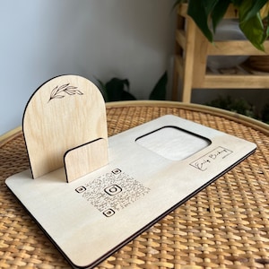 May include: Wooden phone stand with a cutout for a phone and a small shelf for a photo. The stand has a QR code and the text "Emily Baker" on it.