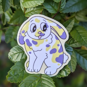 May include: A sticker of a cartoon rabbit with yellow and purple spots. The rabbit has a white body, big eyes, and a pink tongue sticking out.