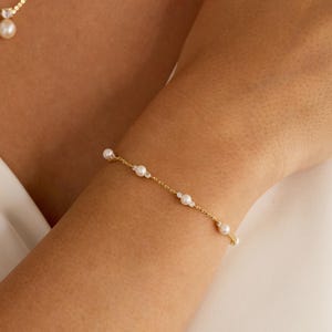 May include: A delicate gold chain bracelet featuring evenly spaced small white pearls and tiny clear crystals. A matching gold necklace with a single pearl pendant is visible at the neckline. The jewelry is worn on a wrist and around the neck.