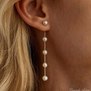 Puede incluir: Un par de pendientes largos de perlas con un solo pendiente de perla en la otra oreja. Los pendientes están en una cadena dorada con varias perlas.