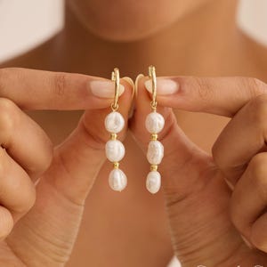 May include: Gold hoop earrings with three white, irregularly shaped pearls suspended from each hoop. The earrings are held up against a blurred background. The earrings have a delicate and elegant design.