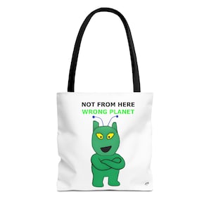 Puede incluir: Bolso tote blanco con asas negras. Presenta un gráfico de un alienígena verde con ojos amarillos y brazos cruzados, y el texto "NOT FROM HERE WRONG PLANET". El bolso está diseñado para transportar objetos.