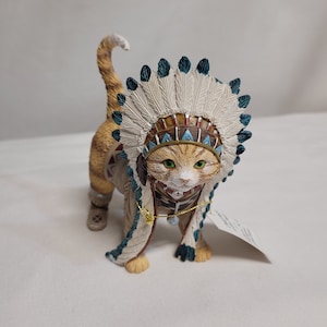 以下が含まれることがあります： 白、青、茶色の羽が付いたネイティブアメリカンの頭飾りをかぶった、詳細なジンジャーのトラ猫の置物。猫はまた、装飾的なベストとブーツを着用しています。猫は緑色の目をし、好奇心旺盛な表情をしています。