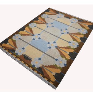 Può includere: Un tappeto rettangolare con un motivo geometrico nei toni dell'azzurro, crema, giallo senape e marrone scuro. Il design presenta motivi floreali simmetrici e un bordo con frange lungo i lati più corti del tessuto intrecciato.