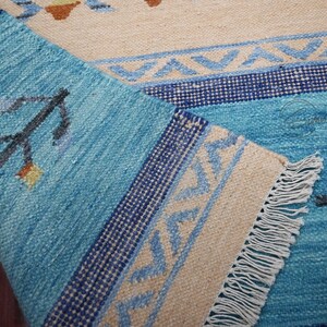 Handwoven kilim rug in blue and beige with tribal geometric patterns; flatweave wool, fringed edge, boho ethnic decor.