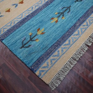 Handwoven kilim rug in blue and beige with tribal geometric motifs and fringe, wool flatweave on dark hardwood.