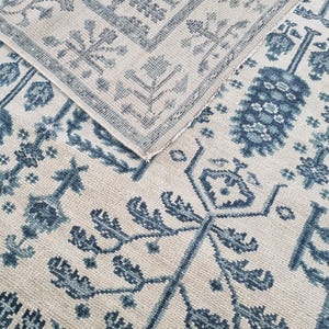 May include: A close-up of a beige rug with a blue floral pattern. The rug has a rectangular shape with a detailed border. The design includes stylized floral and geometric elements, creating a traditional aesthetic. The rug is folded over, revealing the underside.
