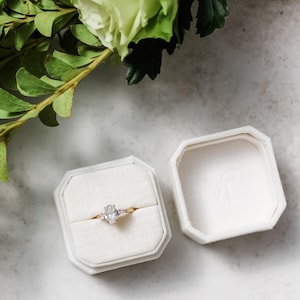 May include: An open, octagonal, cream-coloured ring box displaying a gold ring with an oval-cut diamond and two smaller side stones. The box is on a light surface, with green foliage in the background.