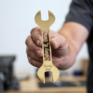 Könnte beinhalten: Ein goldfarbener Schraubenschlüssel mit den eingravierten Texten "Danny R. Brown" und "Thanks Dad". Der Schraubenschlüssel wird von einer Person gehalten, mit unscharfem Hintergrund.
