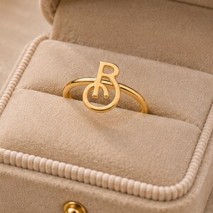 Puede incluir: Un anillo de tono dorado con una letra B estilizada como pieza central, descansando dentro de un joyero de terciopelo beige. El anillo tiene una banda circular delgada y un acabado pulido, presentado en un estuche cuadrado abierto de textura suave.
