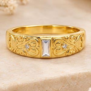May include: A gold band ring featuring a central baguette-cut clear stone flanked by two smaller round-cut clear stones. The metal surface is decorated with intricate embossed scrollwork and heart-shaped patterns across the entire band.
