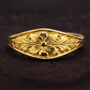May include: A gold ring featuring a central five-petaled flower motif flanked by symmetrical embossed leaf patterns. The band has a polished finish and a tapered design that widens at the center to showcase the detailed floral engraving.