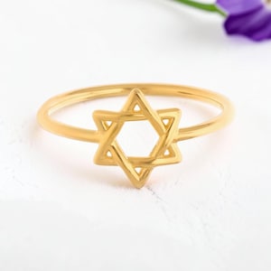 Op de afbeelding: Een gouden ring met een Davidster-ontwerp als centraal middelpunt. De ringband is dun en cirkelvormig en verbonden met het zespuntige ster-embleem. Het metaal heeft een gepolijste, reflecterende afwerking die er helder en glad uitziet.