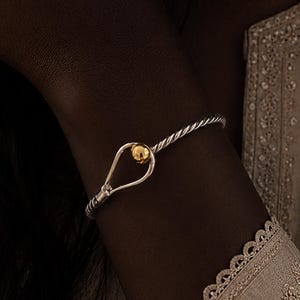 May include: A silver twisted wire bracelet featuring a hook-and-eye style clasp with a single polished gold-tone sphere. The bracelet has a continuous rope-like texture along the band and a teardrop-shaped opening at the centre closure point.