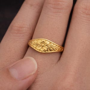 May include: A gold ring with a floral pattern. The ring has an oval face with an embossed flower and leaf design. The ring is displayed on a finger, highlighting the detailed craftsmanship of the jewellery.