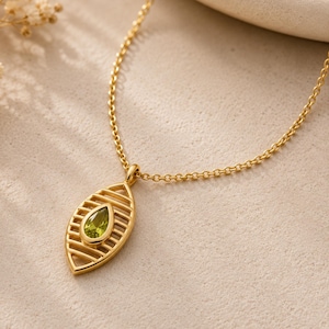 Puede incluir: Un collar de tono dorado con un colgante en forma de marquesa que presenta recortes horizontales y una piedra preciosa de color verde claro en forma de pera en el centro. El colgante cuelga de una delicada cadena dorada, creando una joya.