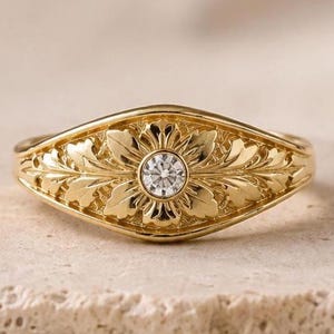 May include: A gold ring featuring a central round clear gemstone set in a bezel. The band is decorated with an embossed leaf pattern that radiates outward from the center stone, creating a detailed botanical design across the front surface.