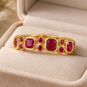 May include: A gold ring featuring a series of alternating rectangular and circular red gemstones set in a band. The ring is displayed inside a beige velvet jewelry box with the text Forever Special and a small heart symbol embossed inside.