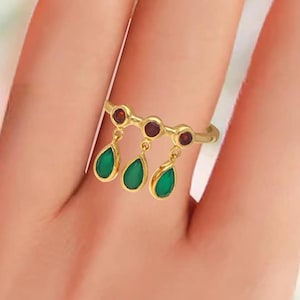 Pode incluir: Anel dourado com três pedras penduradas em forma de gota verde esmeralda e três pedras redondas menores, vermelho escuro. O anel é usado no dedo, mostrando seu design intrincado e cores vibrantes. Uma peça de joalheria única.