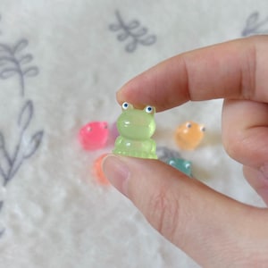 May include: A tiny, translucent green frog figurine with blue eyes is held between fingers. Other colourful frog figurines in pink, orange, and blue are visible in the background. The frog figurines are likely decorative items or charms.