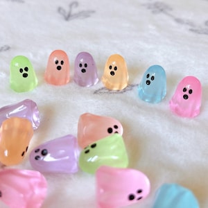 Op de afbeelding: Een verzameling kleurrijke, doorschijnende spookvormige bedels. De bedels zijn in verschillende pastelkleuren, waaronder groen, oranje, paars, geel, blauw en roze. Elke geest heeft drie zwarte stipogen. De bedels liggen op een wit, getextureerd oppervlak.
