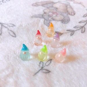 May include: Six miniature gnome figurines with colorful hats in shades of red, orange, purple, green, blue, and pink. The gnomes have clear bodies and white beards, arranged on a soft, white, patterned surface.