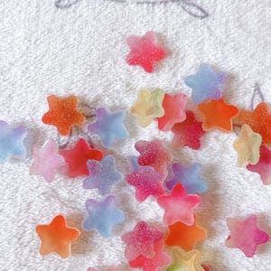Peut inclure: Un ensemble de bonbons en forme d'étoiles multicolores, dans des tons de rouge, orange, rose, bleu et jaune. Les bonbons sont disposés sur une surface blanche et texturée, créant une présentation joyeuse et vibrante. Les bonbons semblent être enrobés de sucre.
