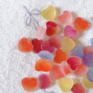 Op de afbeelding: Een verzameling kleurrijke, hartvormige snoepjes in tinten rood, oranje, roze, geel en paars. De snoepjes zijn verspreid op een wit, getextureerd oppervlak, wat een levendige en speelse uitstraling creëert.