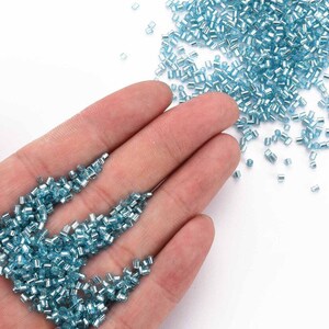 May include: A close-up of a hand holding a pile of small, light blue glass beads. The beads are scattered on a white surface.