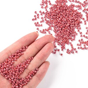 May include: A hand holding a pile of small, round, pink beads. The beads are scattered on a white surface.