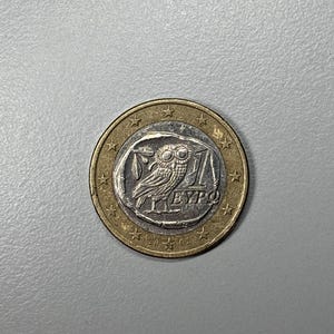 Op de afbeelding: Een euromunt van één euro met een gouden buitenring en een zilveren middenstuk. Het zilveren middenstuk toont een uilontwerp met de tekst "1 EURO" en het jaar "2002". De munt is een symbool van de eurozone.