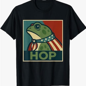 Puede incluir: Camiseta negra con un diseño gráfico de una rana verde con un diseño patriótico. La rana lleva una bufanda con estrellas y rayas, sobre un fondo rojo y azul. La palabra "HOP" está impresa debajo.
