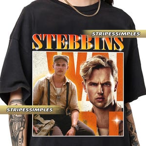 Puede incluir: Camiseta negra con un diseño de cartel de película. El diseño presenta la palabra "STEBBINS" en una fuente degradada naranja y amarilla, y dos hombres con ropa vintage. También es visible la marca "STRIPESSIMPLES".