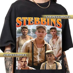 Puede incluir: Camiseta negra con un gráfico de un grupo de hombres y la palabra "STEBBINS" en naranja y blanco. La imagen tiene una estética vintage con rayos y el texto "STRIPESSIMPLES".