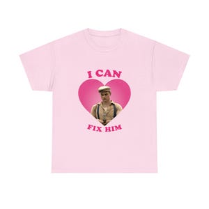May include: A light pink t-shirt with the text "I CAN FIX HIM" in pink lettering. The design features a pink heart shape with a photograph of a man wearing a cap and suspenders.