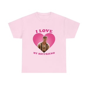 Puede incluir: Camiseta rosa claro con el texto "I LOVE MY BOYFRIEND" en rosa. El diseño presenta un corazón rosa con una foto de un hombre en su interior. La camiseta es de estilo casual.