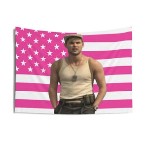 May include: A pink and white American flag tapestry with a person in front of it. The flag has white stars on a pink background and pink and white stripes. The person is wearing a tan tank top, a military-style cap, and dark pants.