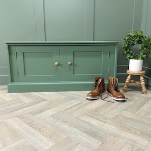 May include: A green cabinet with two doors and wooden handles. Brown leather boots with laces are in front of the cabinet. A small plant in a white pot sits on a wooden stool. The cabinet is 120 cm wide.