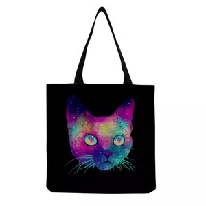 May include: A black tote bag featuring a colourful galaxy cat design. The cat's eyes are bright blue and the galaxy design is in shades of pink, purple, blue and green.