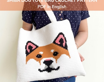 May include: White crochet tote bag featuring a Shiba Inu dog design. The dog's face is rendered in shades of brown, black, and pink. The bag has a rectangular shape with a wide, white strap. Text on the bag reads "SHIBA DOG TOTE BAG CROCHET PATTERN PDF in English".