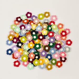 Könnte beinhalten: Eine Sammlung kleiner, gehäkelter Blumen in verschiedenen Farben, darunter Rot, Rosa, Gelb, Grün, Blau und Lila. Jede Blume hat eine kontrastierende Mitte, wodurch ein lebendiges und strukturiertes Aussehen entsteht. Diese handgefertigten floralen Verzierungen sind ideal für Bastelprojekte.