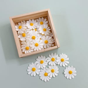 May include: A wooden box filled with white crocheted daisy flowers with yellow centers. The box is sitting on a light green surface. There are also several daisy flowers scattered on the surface.