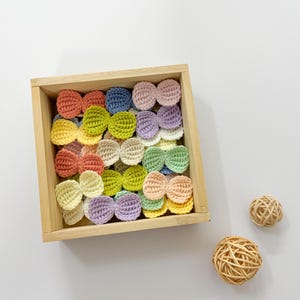 May include: A wooden box filled with crocheted butterfly shapes in various pastel colors, including pink, yellow, blue, and green. The box is sitting on a white surface.
