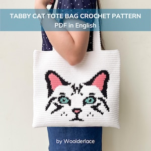 Op de afbeelding: Witte gehaakte tote bag met een tabby kattengezicht ontwerp, roze oren en turquoise ogen. De tas is rechthoekig met twee witte handvatten. De tekst op de tas luidt "TABBY CAT TOTE BAG CROCHET PATTERN PDF in English".